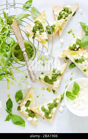 Knusprige Toast (Krostini, Bruschetta) mit Hüttenkäse (Ricotta) und frisch zubereitetem Pesto. Hausgemachte Basilika-Pechto-Soße in einem Glas auf einem hellen Backgroschen Stockfoto