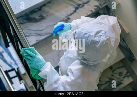 Eine Person, die eine Maske, Handschuhe und einen Sicherheitsanzug trägt, reinigt und desinfiziert eine Tür einer Gemeinschaftswohnung angesichts einer Virus-Pandemie, einer geschützten Safette Stockfoto