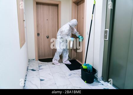 Eine Person, die eine Maske, Handschuhe und einen Sicherheitsanzug trägt, reinigt und desinfiziert eine Tür einer Gemeinschaftswohnung angesichts einer Virus-Pandemie, einer geschützten Safette Stockfoto