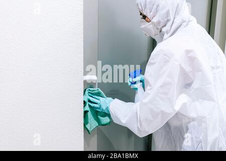 Eine Person, die eine Maske, Handschuhe und einen Sicherheitsanzug trägt, reinigt und desinfiziert eine Tür einer Gemeinschaftswohnung angesichts einer Virus-Pandemie, einer geschützten Safette Stockfoto