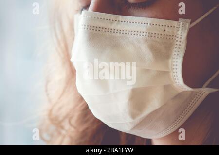 Detail der kaukasischen Frau, die eine weiße medizinische Gesichtsmaske trägt. Frau weint, geschlossene Augen. Fokussieren Sie den vorderen Teil des Gesichts und den verschwommenen Hintergrund. Coronavirus, COVID-19 Quarantäne. Pflegekonzept. Stockfoto