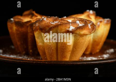 Köstliche, frisch gebackene, saftige Muffins, die mit Puderzucker auf einer Keramikplatte auf schwarzem Grund bestreut werden Stockfoto