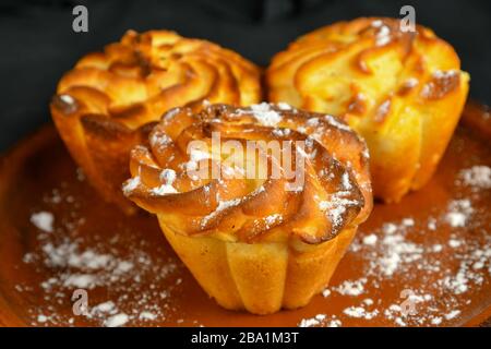 Drei köstliche, frisch gebackene, saftige Muffins, die mit Puderzucker auf einer Keramikplatte bestreut werden Stockfoto