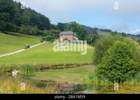 Typische englische Landschaft im Sommer mit einem zentralen Bauernhaus auf einem Hügel mit einer einzelnen Strecke, Teich, Feldern und Schafen unter blauem Himmel Stockfoto