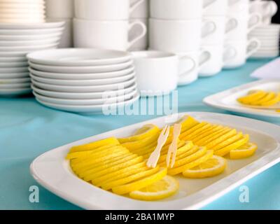 Holzspieße liegen auf einem Stapel dünner Zitronenscheiben, die in einem Stapel ausgelegt sind. Im Hintergrund stehen Stapel weißer Keramikbecher und Untertasse. Tea Party i Stockfoto