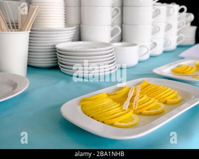 Holzspieße liegen auf einem Stapel dünner Zitronenscheiben, die in einem Stapel ausgelegt sind. Im Hintergrund stehen Stapel weißer Keramikbecher und Untertasse. Tea Party i Stockfoto