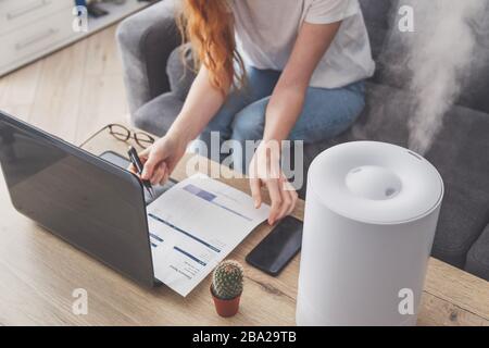 Freiberuflerin verwendet einen Hausbefeuchter am Arbeitsplatz im Heimbüro mit Laptop und Dokumenten Stockfoto