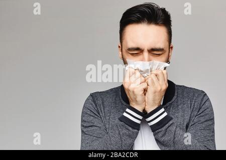 Der junge Mann niest und bedeckt den Mund mit PapierServiette, die auf grauem Hintergrund steht, isoliert. Studio Bild eines jungen Mannes mit Taschentuch. Krank Stockfoto