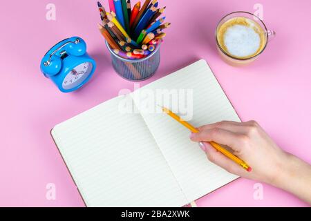 Tablette mit weißem Blatt, daneben eine grüne Pflanze, ein Glas mit Bleistiften. Hand hält einen Stift und schreibt eine Uhr auf das Blatt in einer anderen Hand Stockfoto