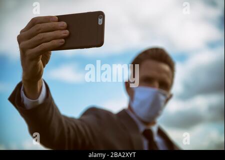Geschäftsmann, der eine schützende, chirurgische Gesichtsmaske trägt, die draußen steht und ein Smartphone selfie nimmt Stockfoto