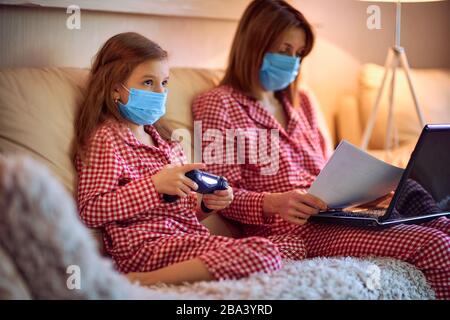 Frau in Pyjamas mit Notizbuch und Papieren, die von zu Hause aus arbeiten und Schutzmaske tragen, während ihr Kind, Tochter Computerkonsolenspiele spielen. Stockfoto