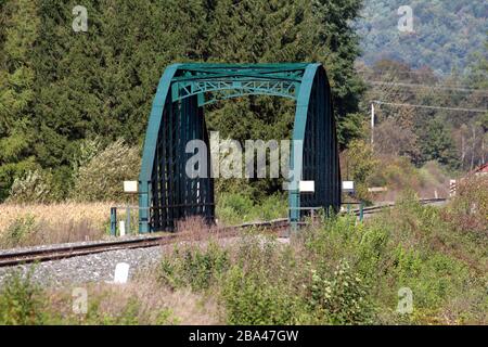 Dunkelgrüne Bahnbrückenkonstruktion aus Metall, die über Bahngleisen und Schotterfundament steigt und mit dichter Waldvegetation und Bäumen umgeben ist Stockfoto