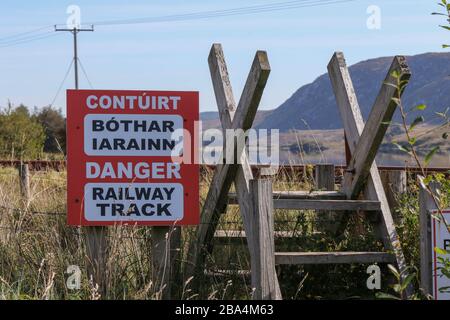 Ein rot-weißes Warnschild in irischer und englischer Sprache neben einem Holzstile an der Fintown Railway Track in einer Gaeltacht Gegend der Grafschaft Donegal, Irland. Stockfoto