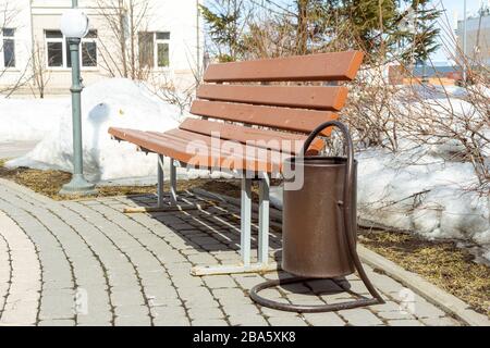 Eine Bank mit einer Urne am Rande des Gehwegs von Blöcken neben einem Busch, der auf einer Rasenfläche wächst, eine Parklampe im Abstand, hinter Bäumen eines Gebäudes Stockfoto