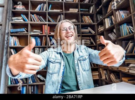 Junge bärtige lächelnde Studentenbloggerin schaut mit den Daumen nach oben. Stockfoto