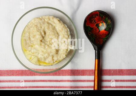 Haferbrei mit Butter in einem Glasbecher und einem Holzlöffel auf einem weißen Tischtuch Stockfoto