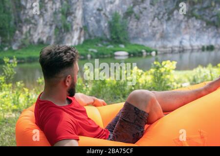 Ein schöner Mann, der sich auf einer aufblasbaren Matratze zurückschlingt, erholsam ist und am schönen Sommertag einen schönen Blick auf den Fluss und die Felsklippen hat. Rückansicht. Reisen Sie an Stockfoto