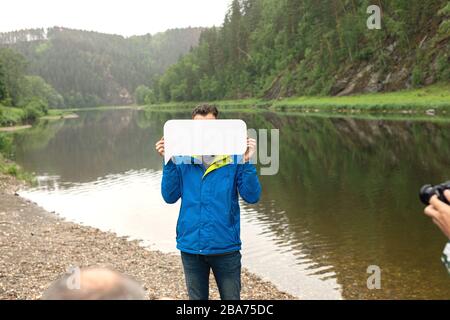 Arbeit des Fotografen mit Modell im Freien. Fotografen, die Nahaufnahmen von einem Mann machen, der einen Teil seines Gesichts hinter einem leeren weißen Bogen im Freien versteckt. Stockfoto