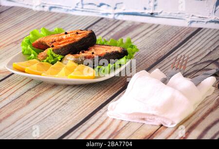 Zwei gegrillte rosafarbene Lachssteaks liegen auf einer weißen Keramikplatte, die mit Zitronenscheiben und einem Salatblatt neben einer weißen Serviette mit Besteck verziert ist, dem WH Stockfoto