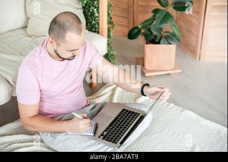 Mann, der online arbeitet, nimmt Notizen auf, die auf dem Boden sitzen Stockfoto