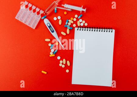 Notepad, eine Vielzahl von Medikamenten, Thermometer, Spritze und Ampullen mit Medikamenten auf rotem Hintergrund. Stockfoto