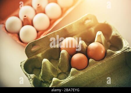 Rohe Eier in weißen und braunen Schalen liegen in roten und grünen Papppaketen, stehen auf einem weißen Tisch und werden durch Sonnenlicht beleuchtet. Stockfoto