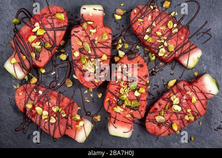 Slices Wassermelone in Eis- oder Popsikelform mit Schokolade und Pistaziennüssen Dekoration auf Schieferplatte, flacher Lay Stockfoto