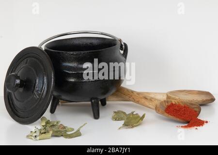 Schwarzer dreibeiniger gusseiserner Topf mit Holzlöffel, Paprika, Senfkörnern und Kalkblättern Stockfoto