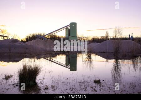 Förderband über Schottergrube bei Sonnenuntergang. Industrielle Landschaft, Gebäude reflektieren im Wasser. Feuchtgebiet Rheinfluss, Deutschland. Stockfoto