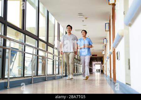 Fröhliche junge asiatische Physiotherapeuten, die im Flur des Pflegeheims spazieren gehen Stockfoto