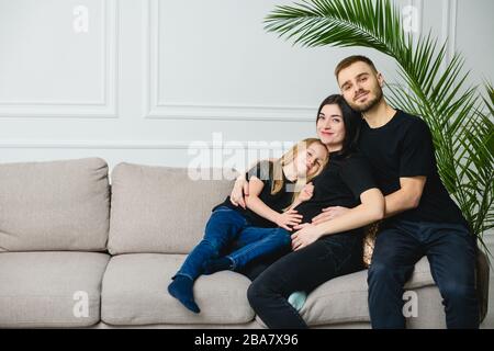 Glückliche Familie im schwarzen Outfit erwartet Baby. Eine junge Familie, Vater, schwangere Mutter und ihre kleine Tochter kuscheln, während sie auf einem Sofa im sitzen Stockfoto