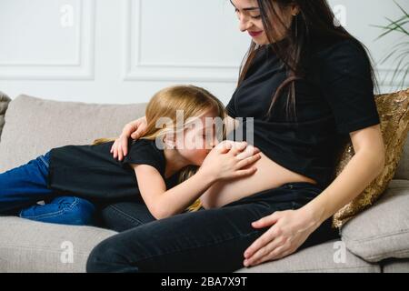 Mädchen küssen Mutter schwangere Bauch auf Couch. Süße schwangere Mutter mit Tochter auf Sofa. Stockfoto