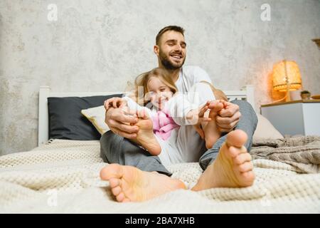 Der junge Vater kitzelt lächelnde Taugher im Bett. Vater und Tochter kitzeln sich gegenseitig die Füße. Stockfoto