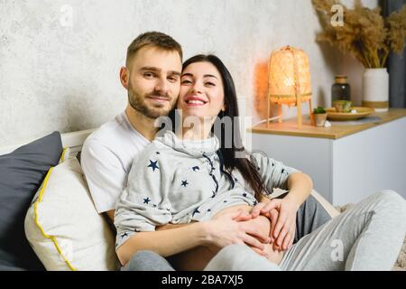 Glückliches junges Paar erwartet Baby zu Hause. Pregant Woman und ihr Mann machen sich mit den Händen über dem Bauch ein Herz. Stockfoto