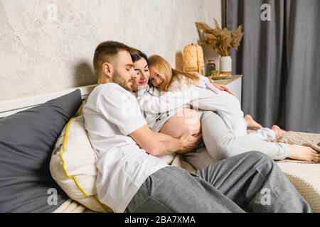 Glückliche Familie im Bett. Glücklicher Vater, Tochter und schwangere Mutter in Pyjamas gekleidet. Mann umarmt eine schwangere Frau, kleine Tochter umarmt Mutter. Stockfoto
