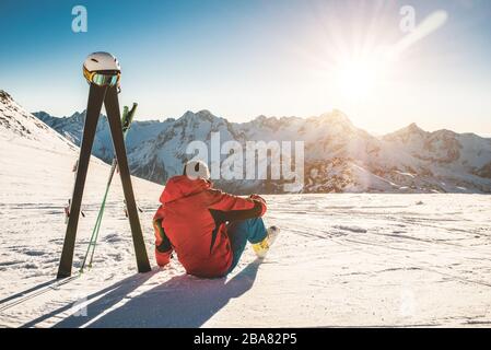 Skifahrersportler, der an sonnigen Tagen in den Schneebergen sitzt - Erwachsener, der den Sonnenuntergang mit Ski-Ausrüstung neben sich genießt - Wintersport- und Urlaubskonzept - Stockfoto