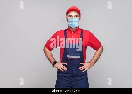 Porträt des jungen Mannes mit chirurgischer medizinischer Maske in Blau insgesamt, rotes T-Shirt und Kappe stehen und halten die Hände auf der Taille und schauen auf Kamera, indoo Stockfoto