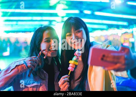 Fröhliche asiatische Mädchen essen Süßigkeiten und nehmen selfie in den Vergnügungspark - junge trendige Freunde, die Spaß am Technologietrend haben - Tech, Freundschaft a Stockfoto