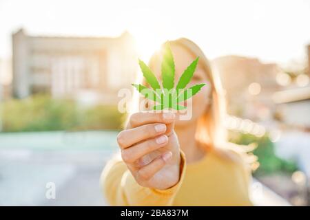 Junges Mädchen, das Marihuanablatt mit Sonnenlicht im Hintergrund hält - Cannabis Medizin, gesunder Lebensstil und Ökologiekonzept - konzentriert sich auf die Hand Stockfoto