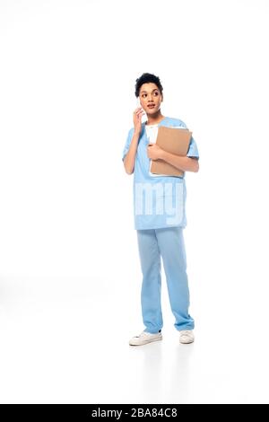 Vollständige Ansicht der afroamerikanischen Krankenschwester mit Papieren, die auf dem Smartphone auf Weiß sprechen Stockfoto