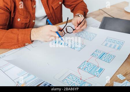 Freigestellte Ansicht des Designers, der Brillen und Marker in der Nähe von Vorlagen für das Benutzererlebnis auf einem Tisch hält, der auf grau isoliert ist Stockfoto