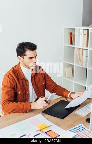 Ansehnlicher ux-Designer, der Smartphone hält, während er mit Website-Skizzen am Tisch im Büro arbeitet Stockfoto