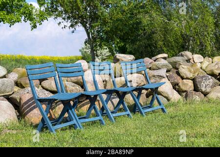 Vier blaue Stühle, in einer Reihe auf der Wiese, vor einer Steinmauer, in der Landschaft, während der Springzeit stehen. War in Mecklenburg zu sehen. Stockfoto
