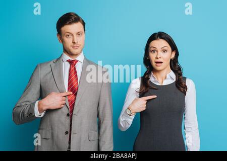 Ein ernsthafter Geschäftsmann und schockierte Geschäftsfrau, die mit den Fingern zeigte, während sie die Kamera auf blauem Hintergrund ansah Stockfoto