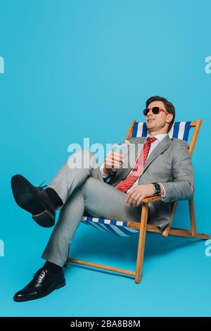 Fröhlicher Geschäftsmann in Sonnenbrille auf dem Liegestuhl mit einem Glas Cocktail auf blauem Hintergrund Stockfoto