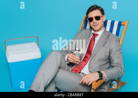 Glücklicher Geschäftsmann in Sonnenbrille auf dem Liegestuhl mit einem Glas Cocktail in der Nähe eines tragbaren Kühlschranks auf blauem Hintergrund Stockfoto