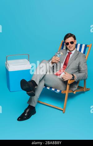 Lächelnder Geschäftsmann in einer Sonnenbrille auf dem Liegestuhl mit einem Glas Cocktail in der Nähe eines tragbaren Kühlschranks auf blauem Hintergrund Stockfoto