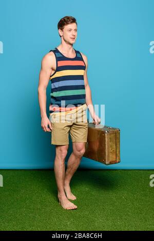 Schöner Mann in gestreifter Singlet und Shorts, die Vintage-Koffer auf blauem Hintergrund halten Stockfoto