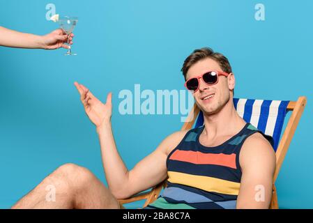 3/4-Blick auf die Frau, die dem Mann ein Glas Cocktail in einer Sonnenbrille auf dem Liegestuhl auf blauem Hintergrund gibt Stockfoto