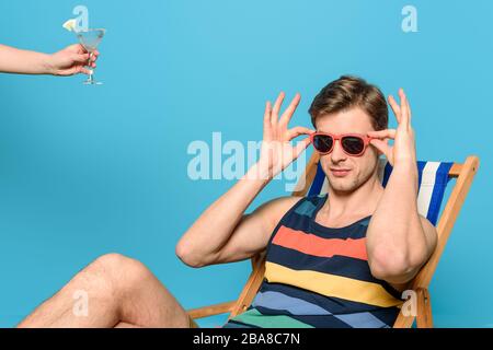 3/4-Ansicht der Frau, die einem Mann, der im Liegestuhl sitzt und eine Sonnenbrille auf blauem Hintergrund berührt, ein Glas Cocktail gibt Stockfoto
